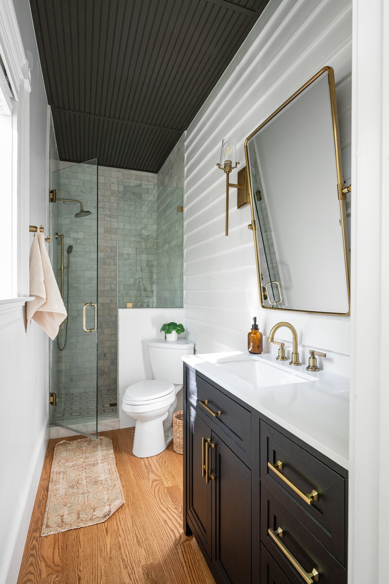 Narrow bathroom inside a Norcross, GA, home with a glass-enclosed marble shower, dark wood vanity, brass fixtures, white shiplap walls, and warm hardwood flooring.