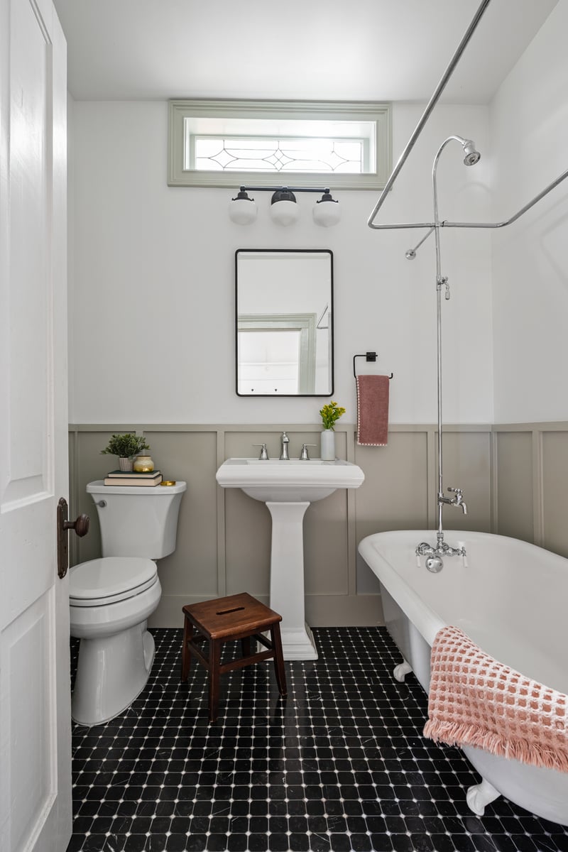 Traditional bathroom inside a Norcross, GA, home  with a clawfoot tub and exposed shower, pedestal sink, black-and-white tile floor, and muted gray wainscoting.