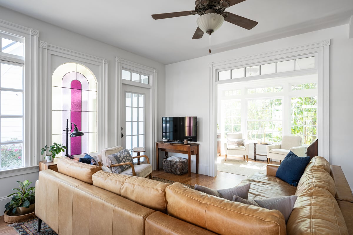 Bright living room inside a Norcross, GA, home with a tan leather sectional, arched stained-glass window, ceiling fan, and an adjoining sunroom with two armchairs.