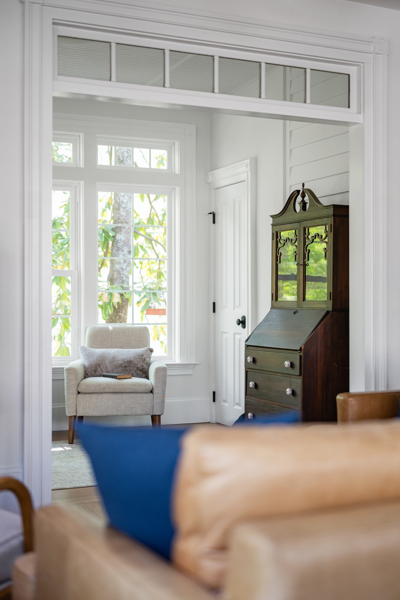 View through a trimmed doorway into a bright sitting area inside a Norcross, GA, home with an upholstered armchair, large windows, and an antique secretary desk.